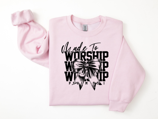 A light pink crewneck sweatshirt with a black graphic print on the front reading 'Made To Worship' and 'PSALM 95:1' around a bow tie illustration, displayed flat on a white background.