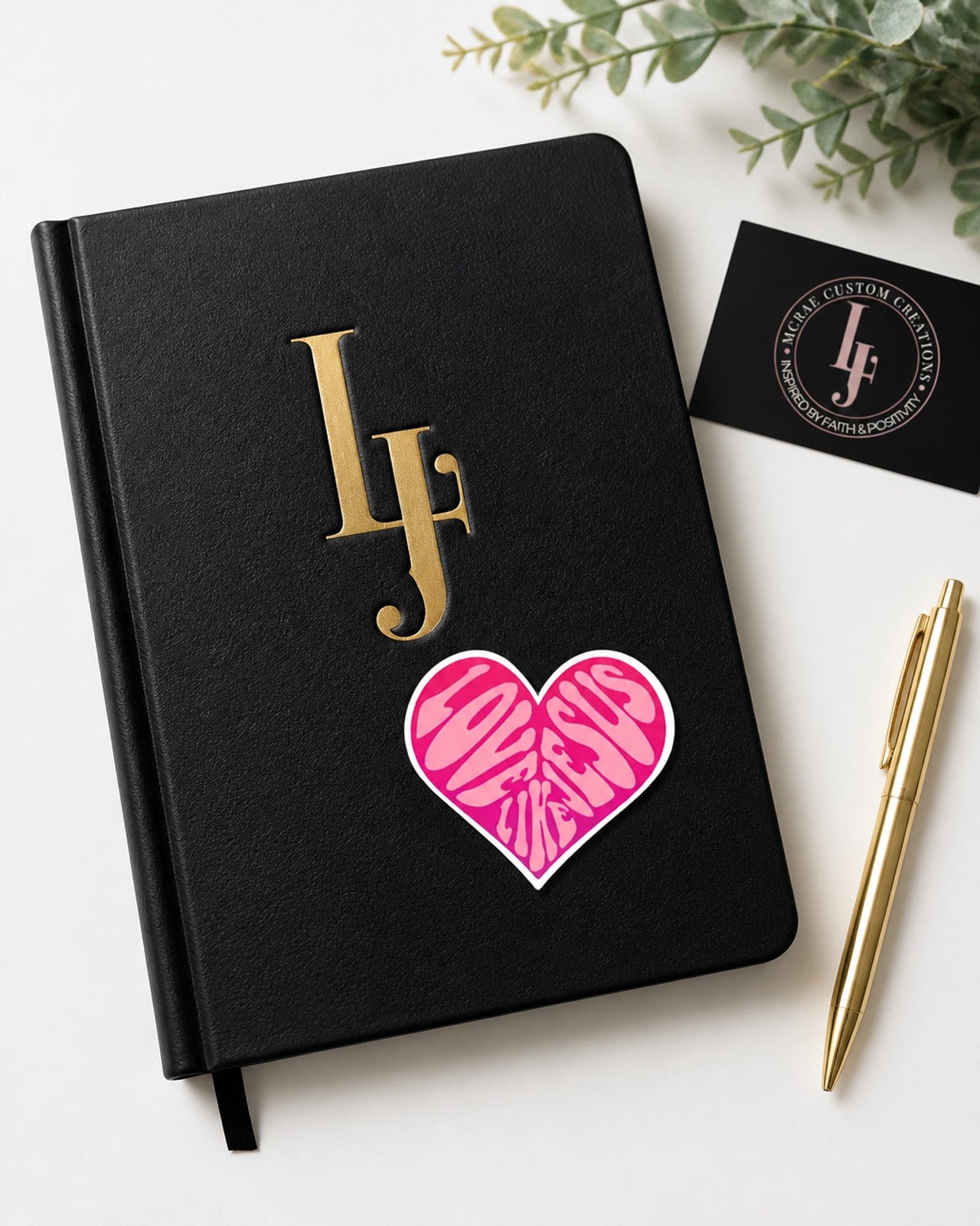 Black notebook with gold initials and pink heart design, placed on a white surface with a pen and business card.